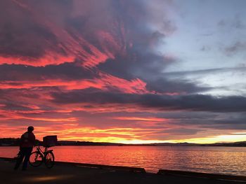 Silhouette man with bicycle standing at beach against dramatic sky during sunset