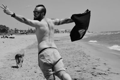 Rear view of shirtless man with arms outstretched standing on beach