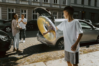 Son playing with inflatable ring standing by family at roadside on sunny day