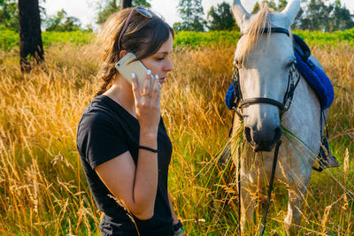 Young woman using phone while standing with horse against sky