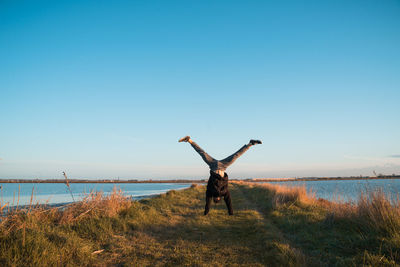 Man doing handstand on field by lake against clear sky during sunset