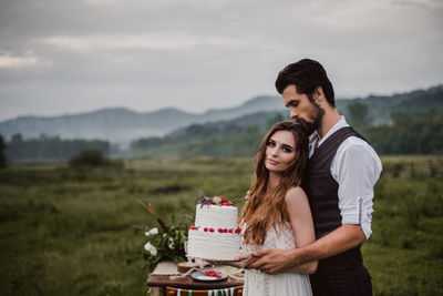 Portrait of bride with bridegroom holding wedding cake on field