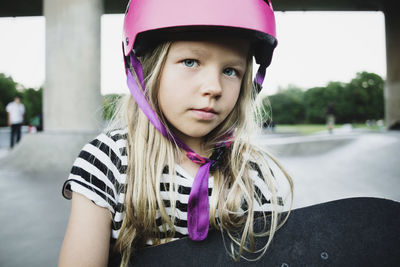 Portrait of girl wearing pink helmet holding skateboard at park