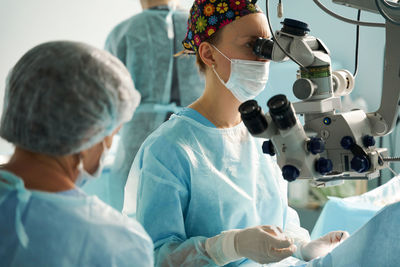 Adult female doctor in sterile mask and ornamental medical cap looking through surgical microscope against crop coworker in hospital