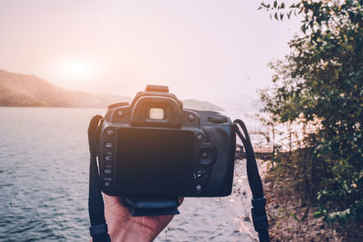 Cropped hand holding camera by lake during sunset