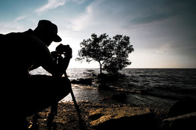 Silhouette person photographing by sea against sky during sunset