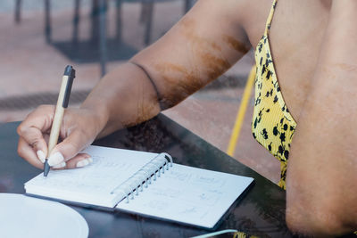 Crop view of black self employed woman sitting at table in cafe and working remotely while writing in notepad