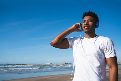 Smiling man listening music while standing at beach against blue sky