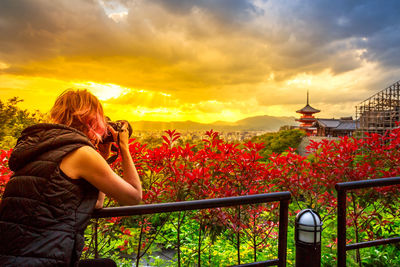 Woman photographing by railing against sky during sunset