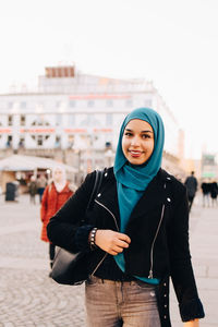 Portrait of smiling young muslim woman wearing hijab walking on footpath in city