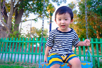 Portrait of cute boy sitting on swing against fence 