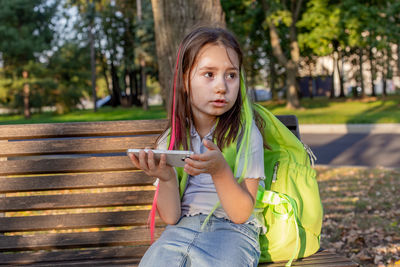 Young girl sitting on park bench with smartphone and backpack on a sunny day