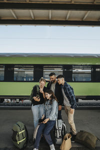 Happy woman sharing smart phone with family standing in front of train at railroad station