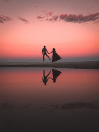 Silhouette couple jumping with reflection in sea against orange sky during sunset