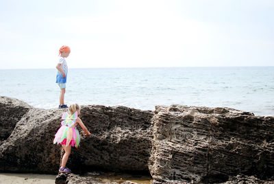 Rear view of two kids standing at rocks beach against sky