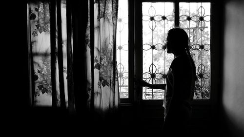 Side view of silhouette teenage girl looking through window at home