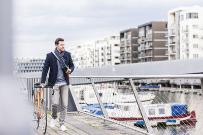 Businessman pushing bicycle in the city, while using smartphone and earphones