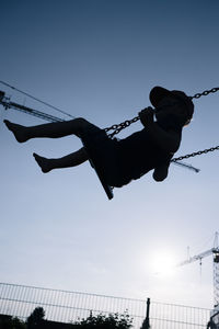 Low angle view of silhouette boy enjoying swing against clear blue sky