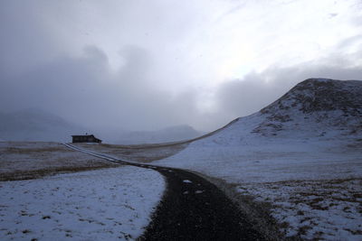 Narrow road by snow covered field against sky