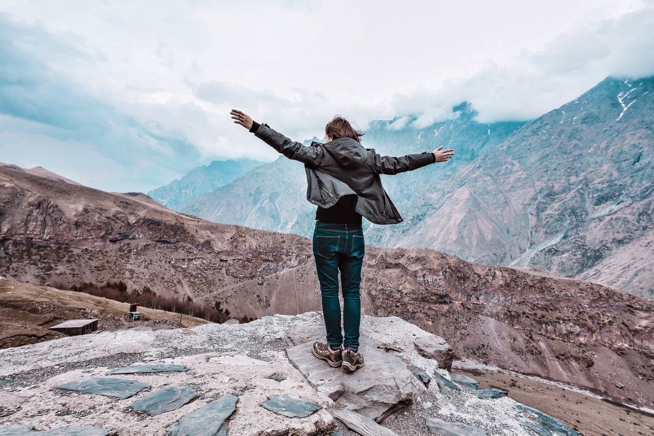 mountain, real people, human arm, one person, standing, arms outstretched, limb, sky, scenics - nature, leisure activity, beauty in nature, full length, lifestyles, mountain range, cloud - sky, casual clothing, non-urban scene, tranquility, tranquil scene, arms raised, outdoors, freedom