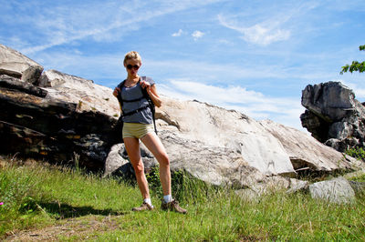 Portrait of female hiker wearing sunglasses standing on grassy field amidst rock formation against sky