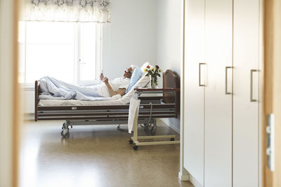 Side view of senior man using smart phone while reclining on bed in hospital ward