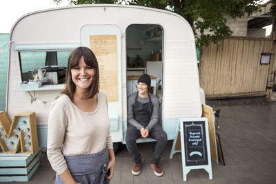 Portrait of happy female owner standing on street while coworker sitting in food truck