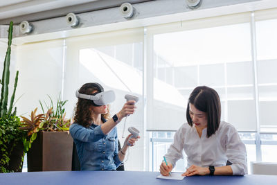 Wide angle view on two women sitting at the table in the office with vr helmet