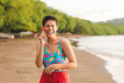 Cheerful young latin american woman with short hairstyle talking on mobile phone while standing with eyes closed near waving sea in summer day