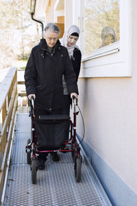 Female home caregiver helping senior woman with walking frame through passage