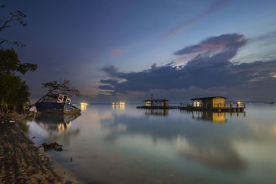 Shipwreck and stilt house on lake against cloudy sky at dusk