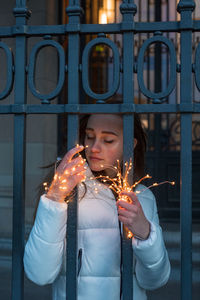 Woman holding illuminated lighting equipment while leaning on gate