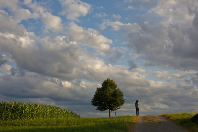 Rear view of woman with hand raised standing on field against cloudy sky during sunset