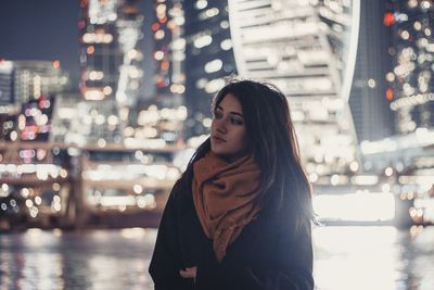 Young woman looking away while standing against illuminated buildings at night