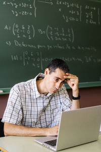 Stressed math teacher sitting at table with laptop in classroom