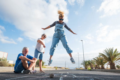 Low angle view of woman jumping during hopscotch with senior couple on footpath against sky