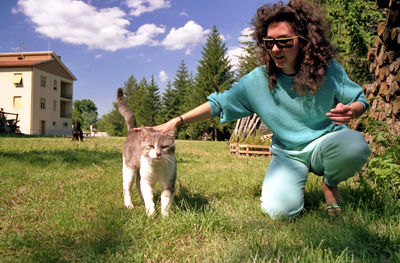 Full length of woman crouching with cat sitting on grass against sky