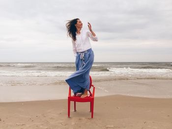 Beautiful woman standing on chair at beach against cloudy sky