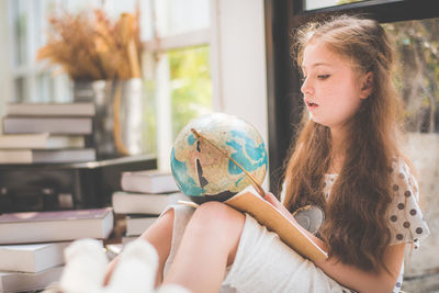 Cute girl looking at globe while sitting against window at home