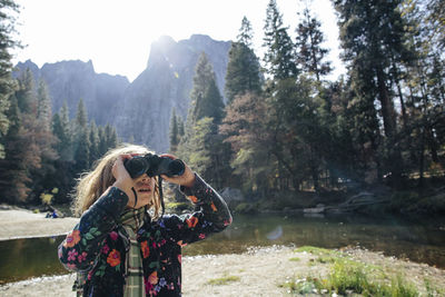 Girl looking through binoculars while standing on lakeshore in forest at yosemite national park during sunny day