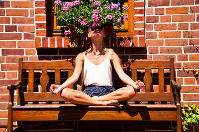 Young woman sitting on bench against brick wall during sunny day