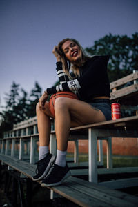 Portrait of smiling young woman sitting on wood against sky