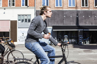 Profile shot of young man holding burger while riding bicycle on city street