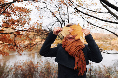 A woman in a coat and scarf in an autumn park covers her face with maple leaves