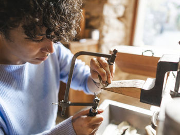 Crop focused hispanic female goldsmith master using manual fretsaw while working with jewelry in light studio