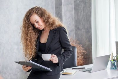 Businesswoman holding coffee cup while reading paper in office