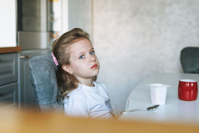 Unhappy little girl with long hair in home clothes with yogurt sitting at dinning table in kitchen 