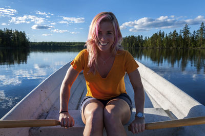 Portrait of smiling young woman sitting by lake against sky