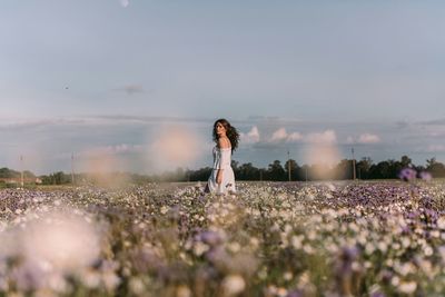 Side view of young woman standing amidst flowers against sky during sunset