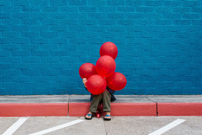 Boy with red balloons sitting on sidewalk against blue wall